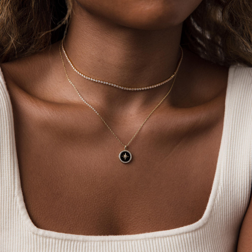 A woman wears two gold necklaces, including the Onyx Star Pendant Necklace in 18K Gold, paired with a white ribbed tank top, shown from neck to upper chest.