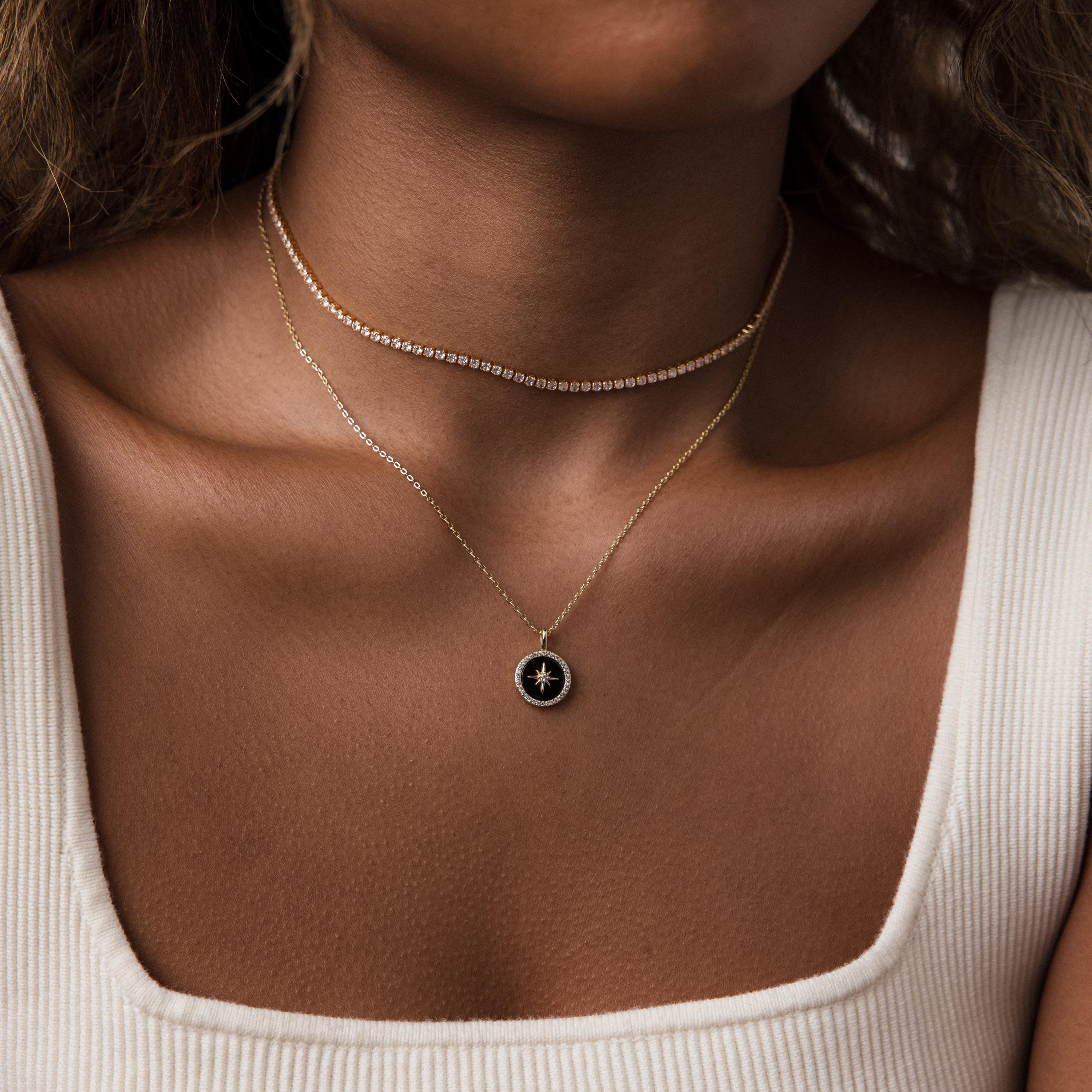 A woman wears two gold necklaces, including the Onyx Star Pendant Necklace in 18K Gold, paired with a white ribbed tank top, shown from neck to upper chest.