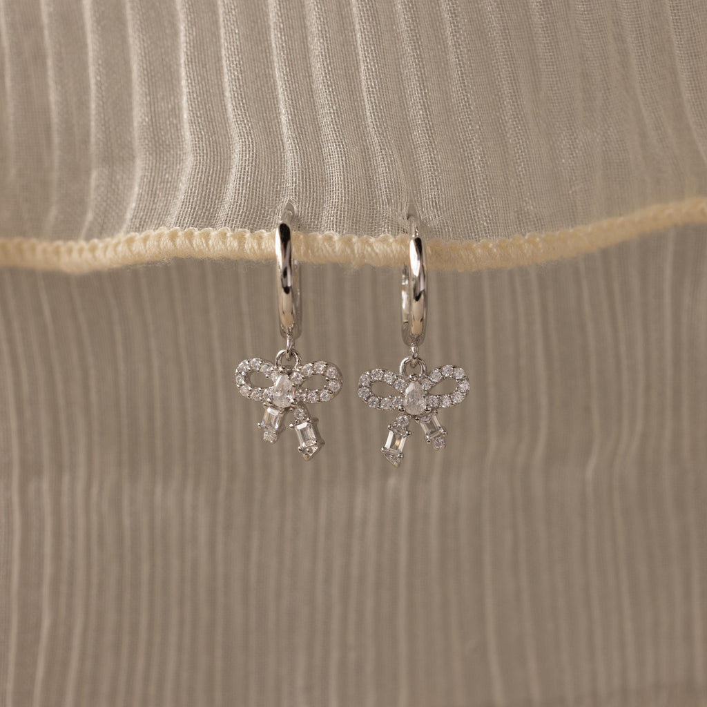 Flatlay of our Pave Bow Charm Hoops
 in Silver on a beige fabric background