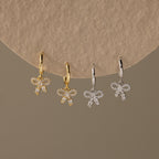 2 pairs of our Pave Bow Charm Hoops earrings in gold and silver on a beige background