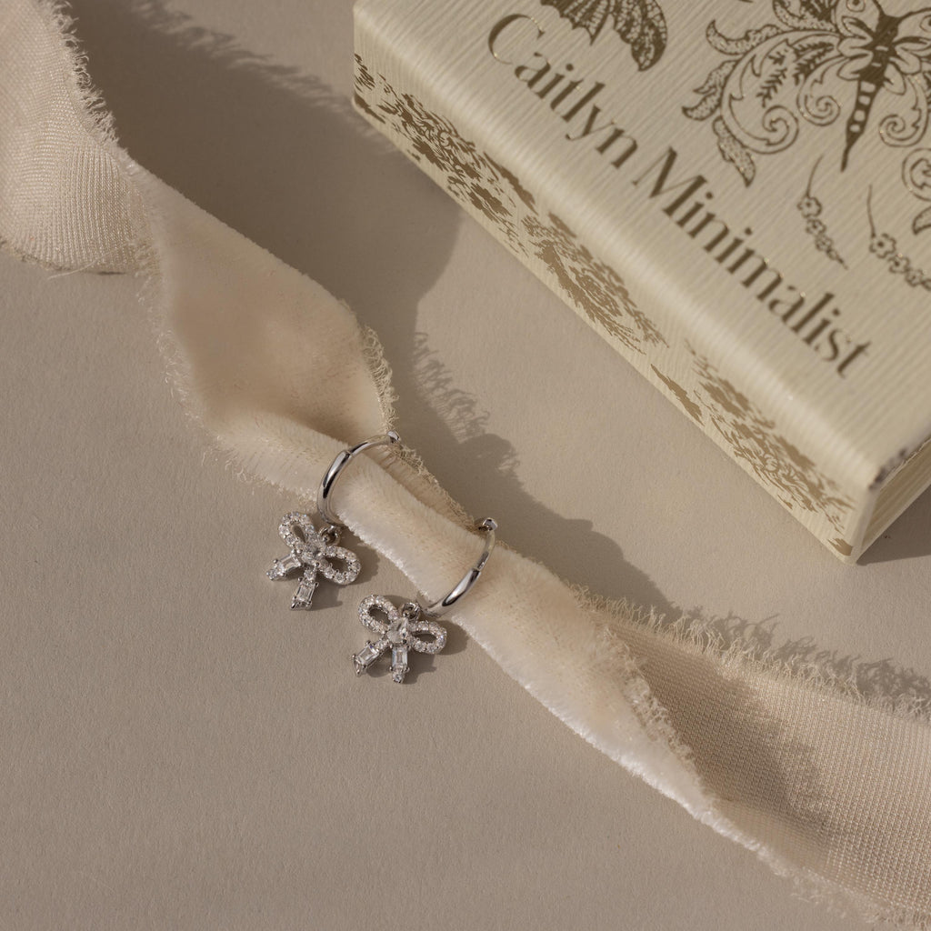 Flatlay of our Pave Bow Charm Hoops in Silver against a beige ribbon with a Caitlyn Minimalist box in the background