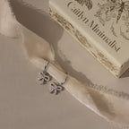 Flatlay of our Pave Bow Charm Hoops in Silver against a beige ribbon with a Caitlyn Minimalist box in the background