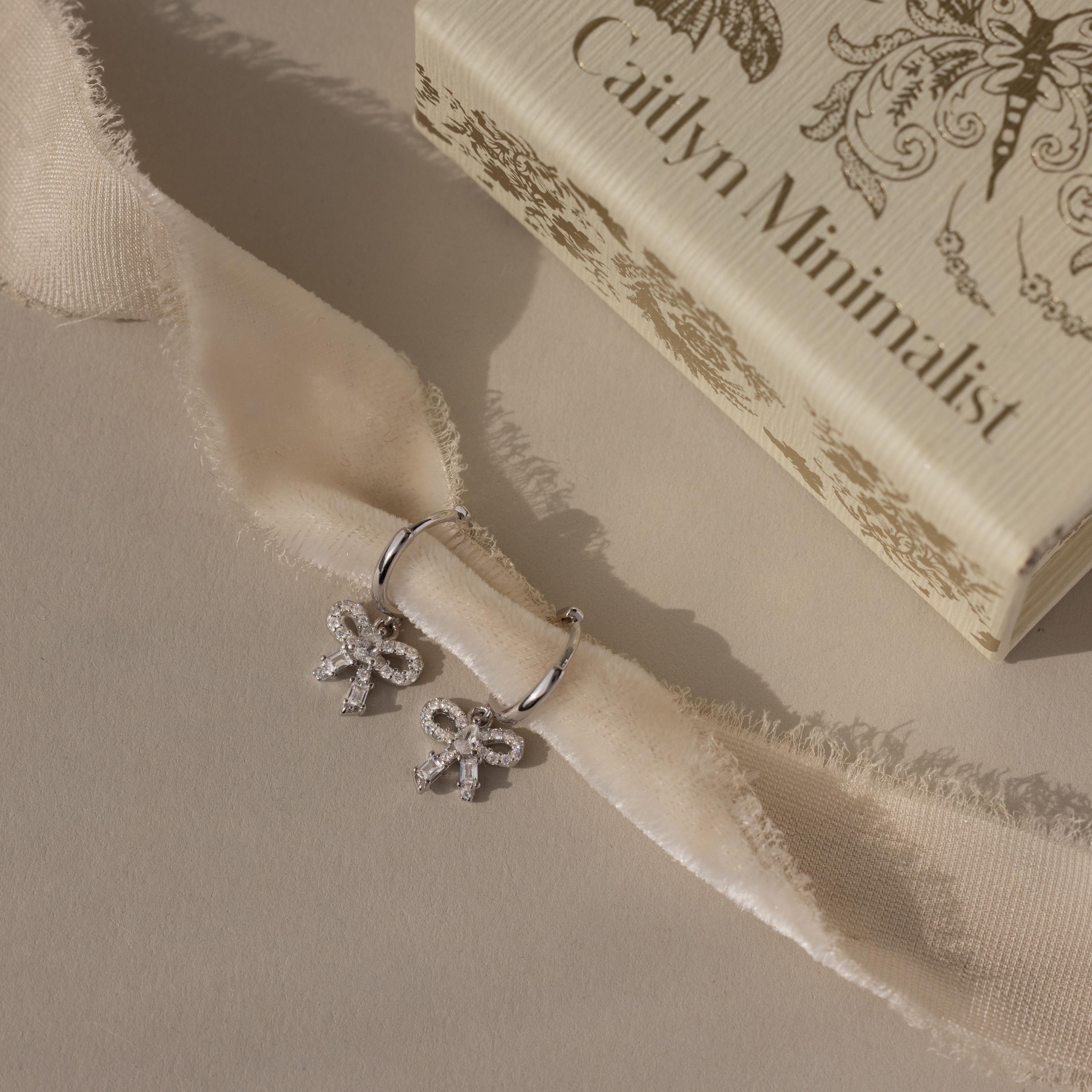 Flatlay of our Pave Bow Charm Hoops in Silver against a beige ribbon with a Caitlyn Minimalist box in the background