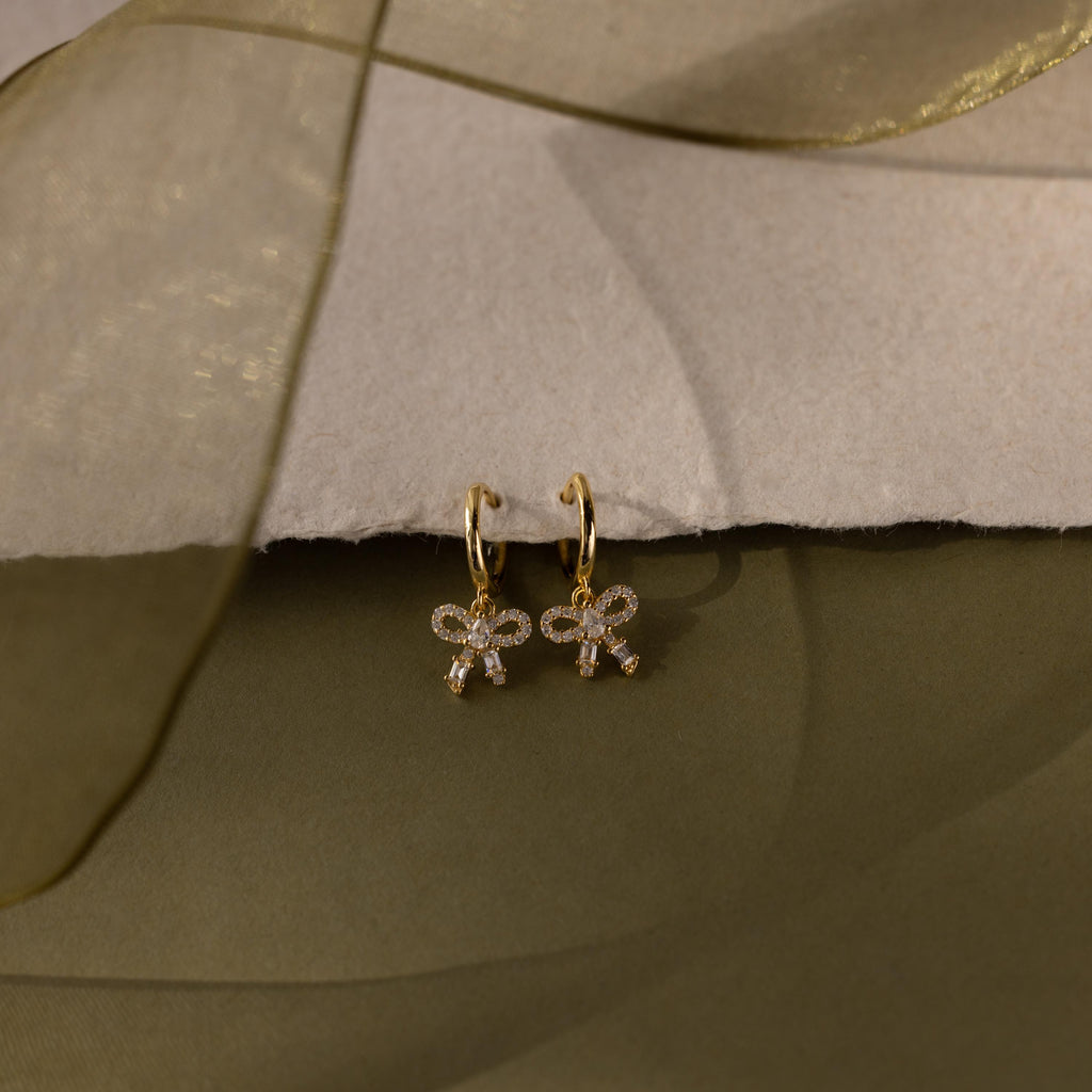 Flatlay of our Pave Bow Charm Hoops earrings in Gold on a soft fabric background