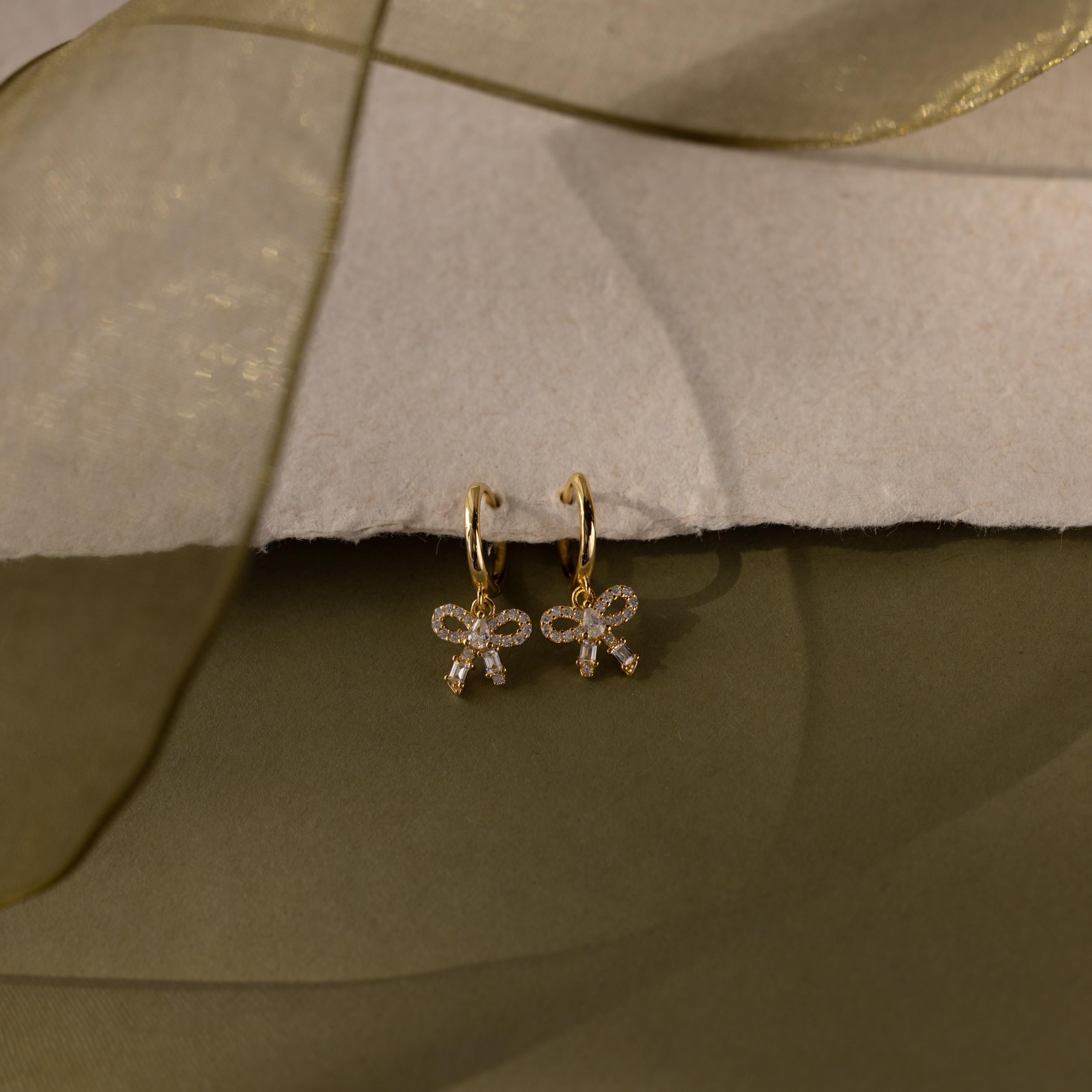 Flatlay of our Pave Bow Charm Hoops earrings in Gold on a soft fabric background