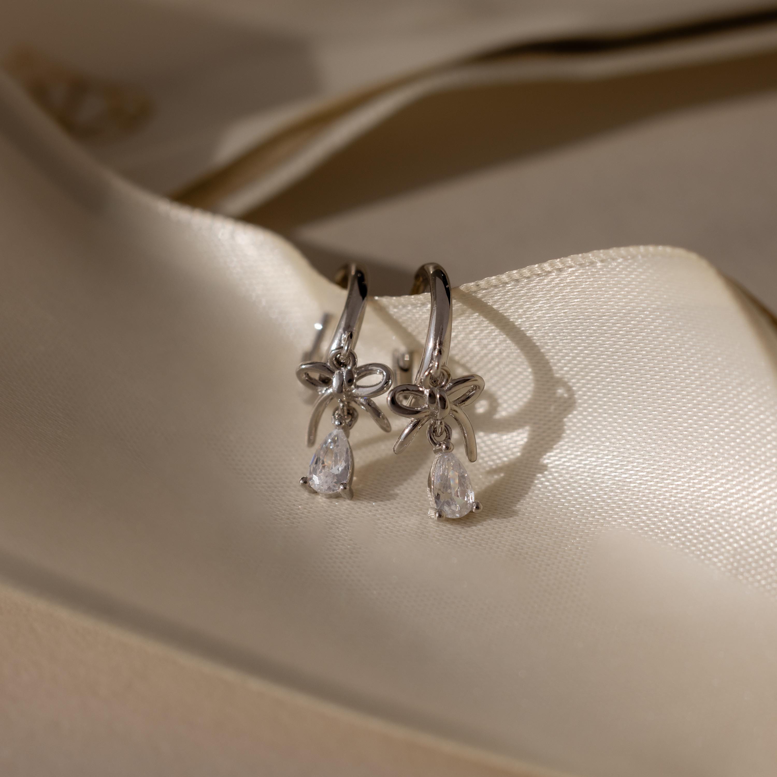 Flatlay of our Teardrop Ribbon Charm Hoops in Silver earrings on a beige fabric background