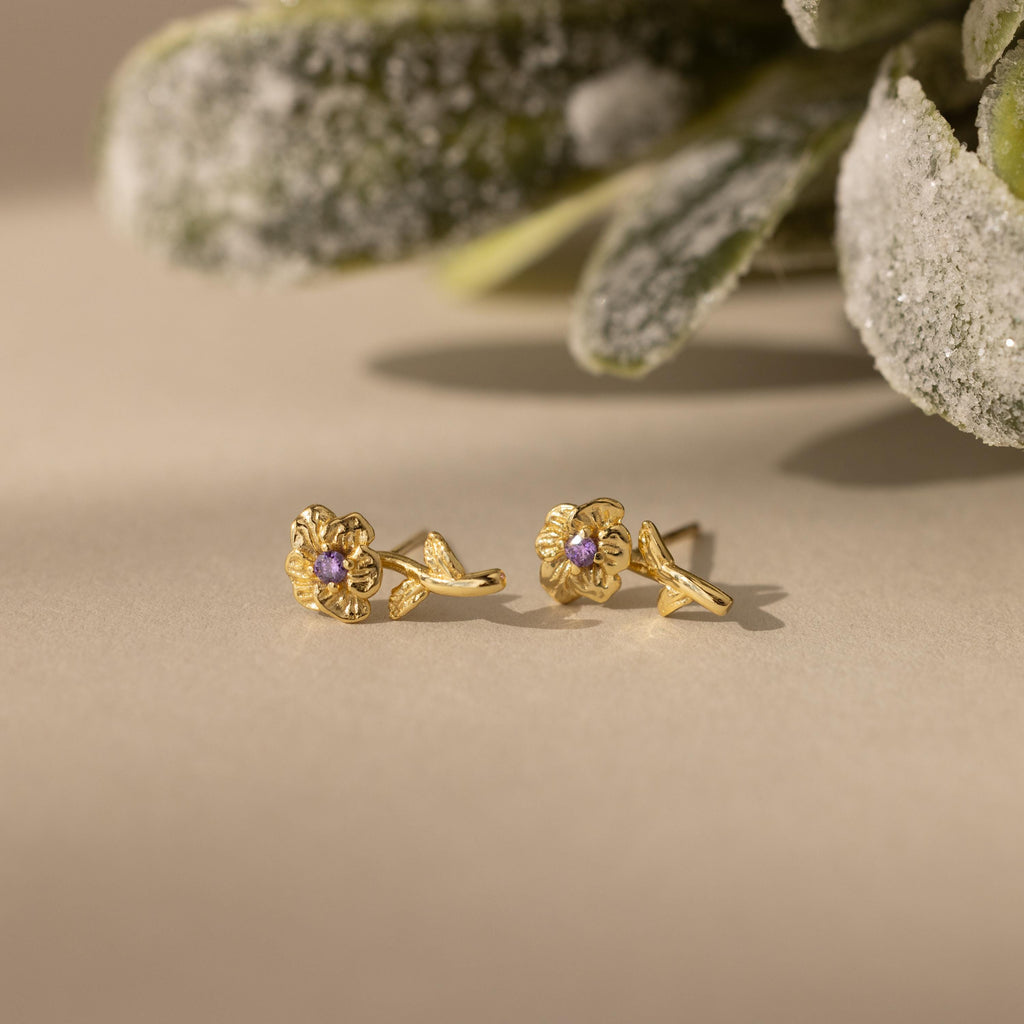 Birth Flower Stud Earrings with Amythest stones on a beige surface with green foliage in the background