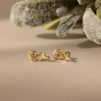 Birth Flower Stud Earrings with Amythest stones on a beige surface with green foliage in the background