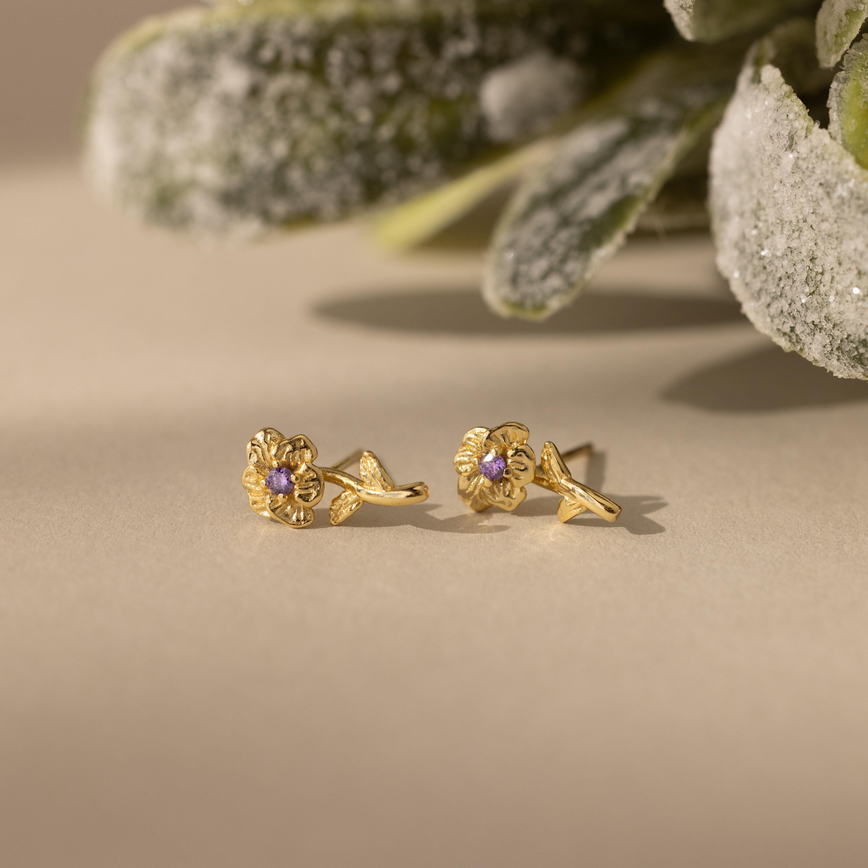 Birth Flower Stud Earrings with Amythest stones on a beige surface with green foliage in the background