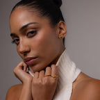 Woman wearing love knot studs with a white turtle neck and gold ring stack against a neutral background