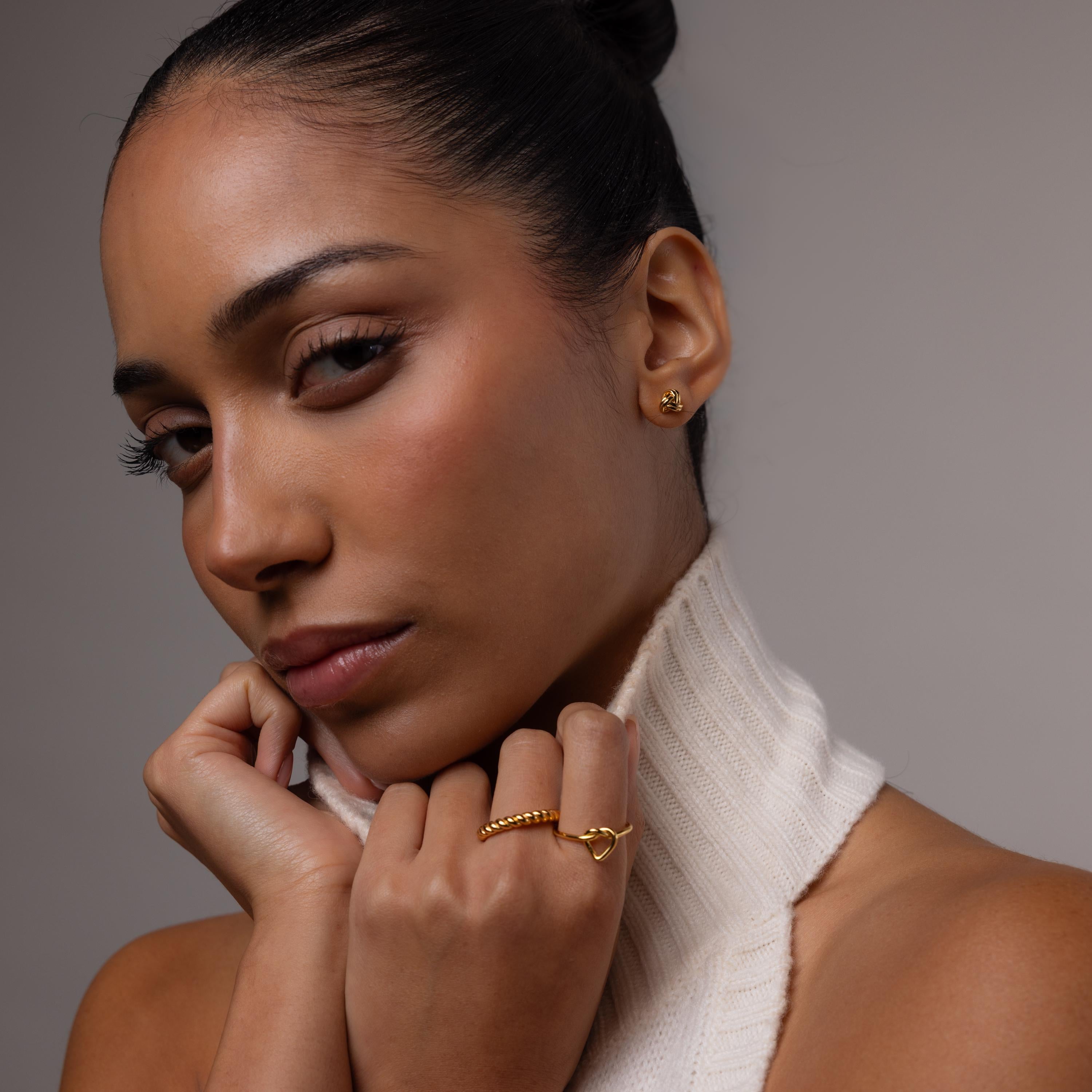 Woman wearing love knot studs with a white turtle neck and gold ring stack against a neutral background