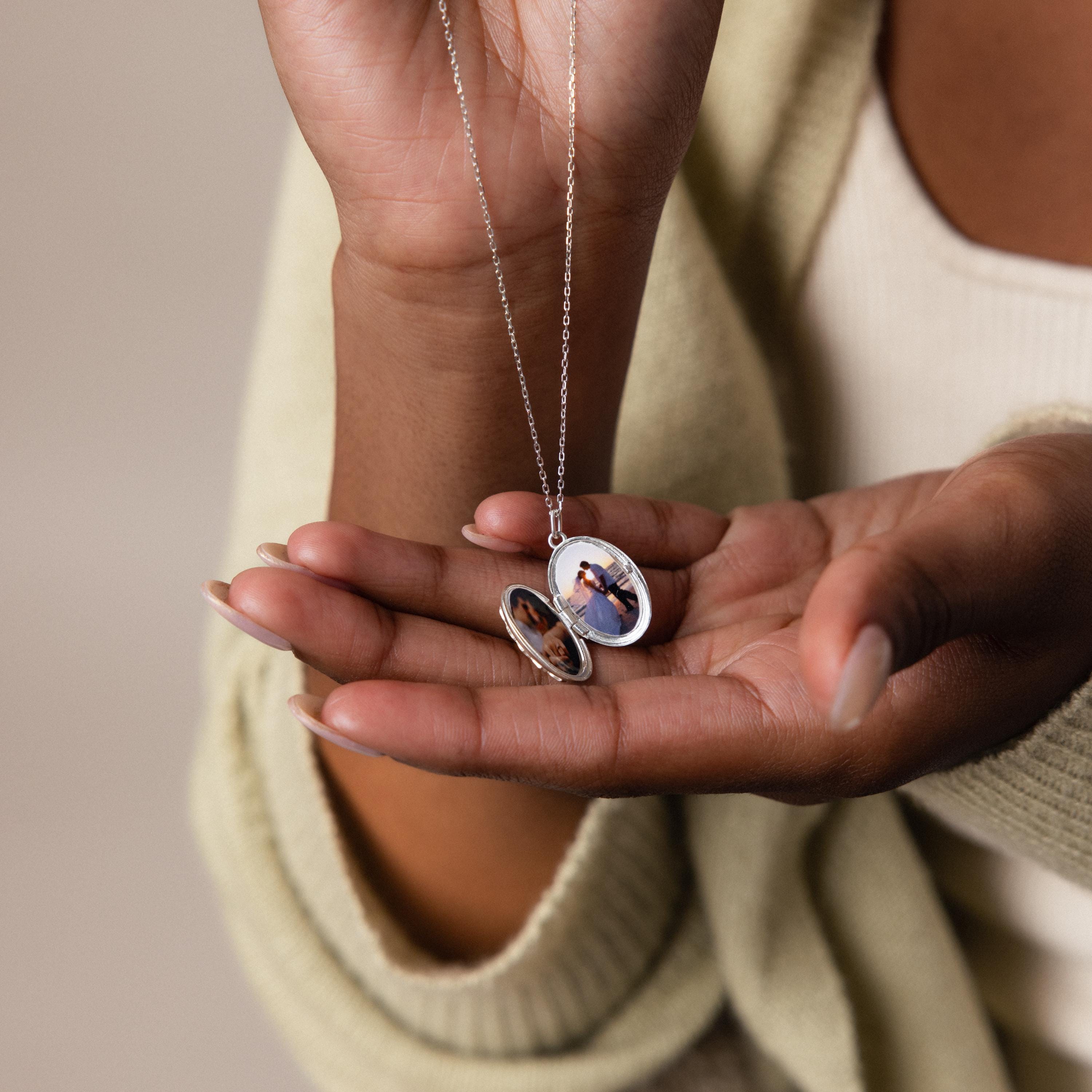 A person holds the Floral Handwriting Locket Necklace in Sterling Silver open, showing a small photo inside along with their hands and part of their outfit.
