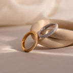 A gold and a silver Skinny Pave Stacking Ring, both featuring diamond-studded pave stones, are displayed on a smooth, curved beige surface.