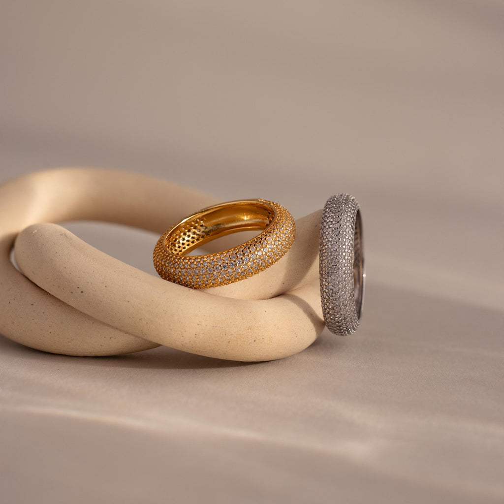 The Bold Pave Stacking Ring in silver rests alongside a gold wedding band on intertwined beige ceramic shapes against a soft backdrop.
