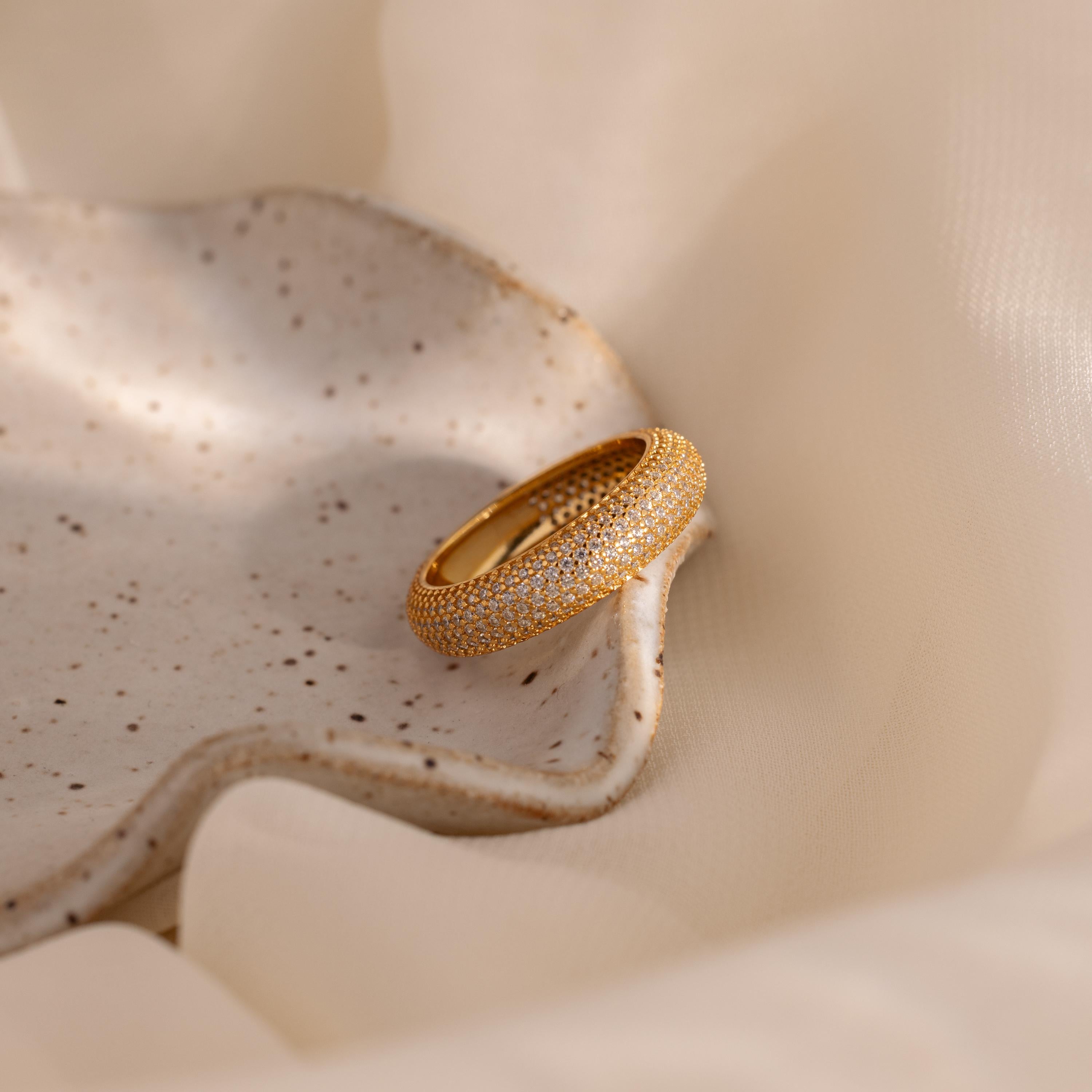 The Bold Pave Stacking Ring, adorned with small white stones, rests on a speckled ceramic dish set against a soft beige fabric background.