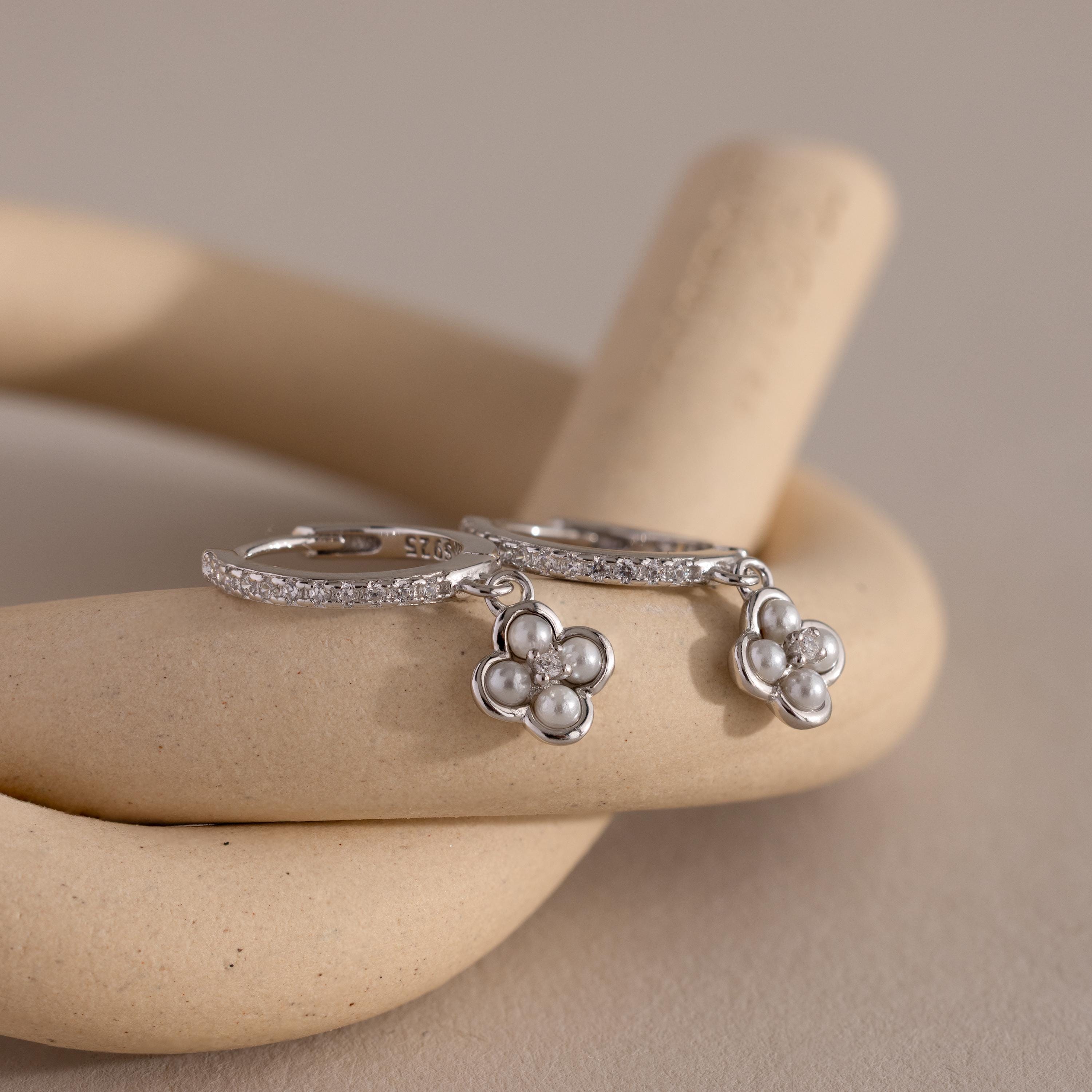 The Pave Rounded Pearl Flower Hoops feature delicate floral charms and pearls, resting on a curved beige display—elegant earrings perfect for weddings and any special occasion.