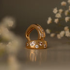 The Diamond Confetti Signet Ring rests on a reflective surface, its gold shimmering amid blurred white flowers.