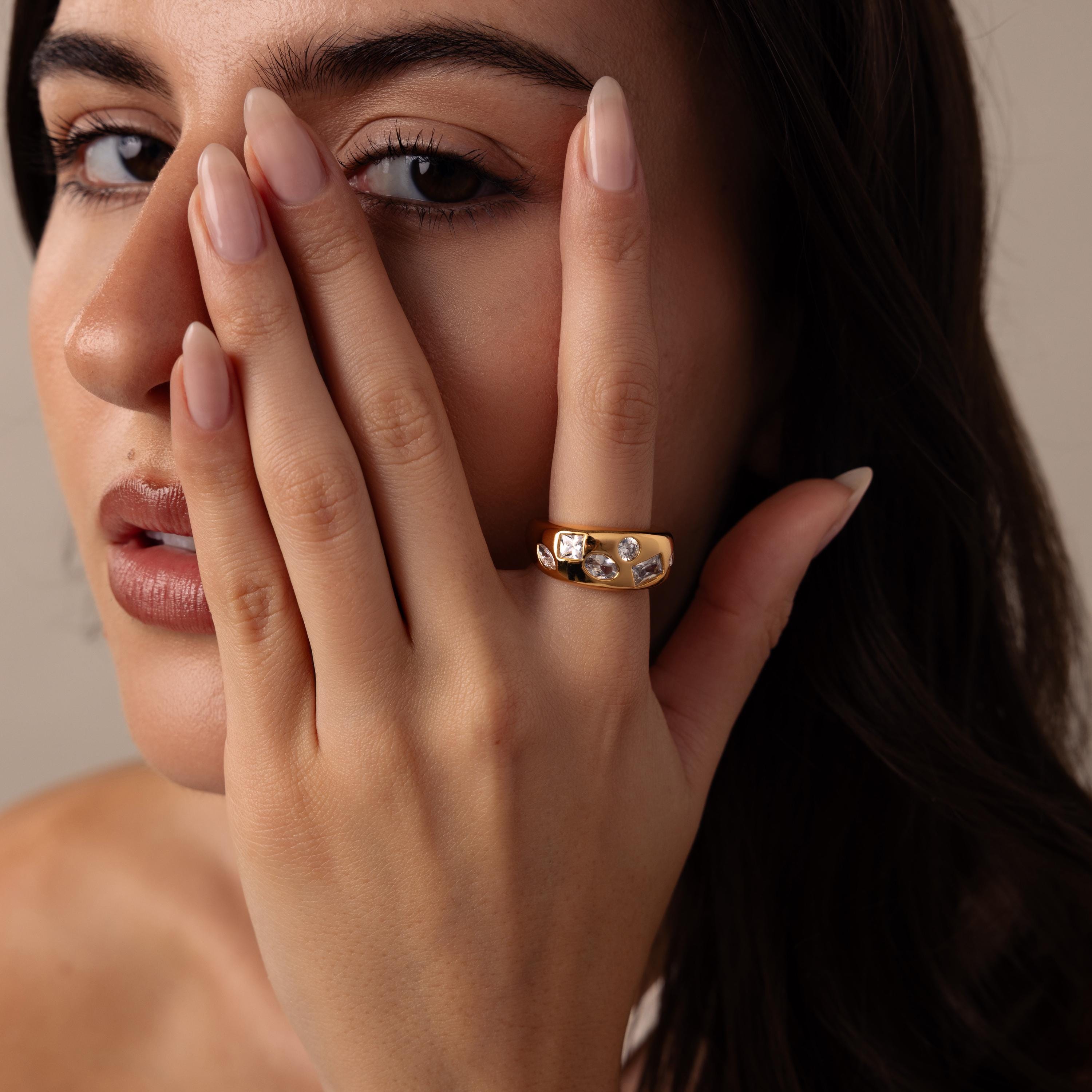 A woman with long nails covers half her face, showcasing the Diamond Confetti Signet Ring on her finger—its modern design adorned with sparkling gemstones.
