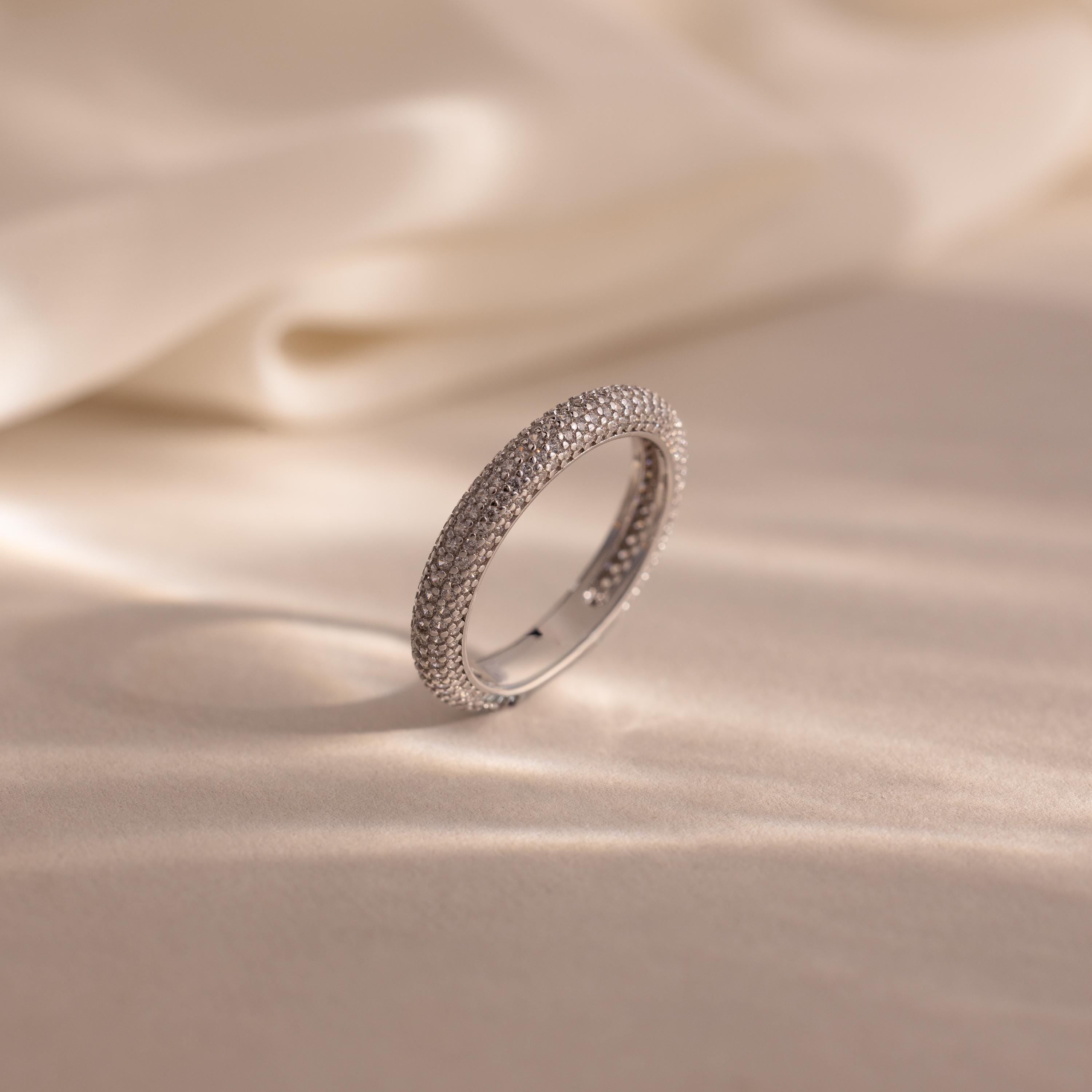 The Skinny Pave Stacking Ring stands upright on a soft beige surface, its sparkling pave stones glimmering with elegance against a backdrop of draped fabric.