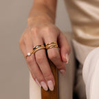 A hand with manicured nails wears the Skinny Pave Stacking Ring, featuring sparkling pave stones, along with several gold rings, gently resting on a wooden armrest.