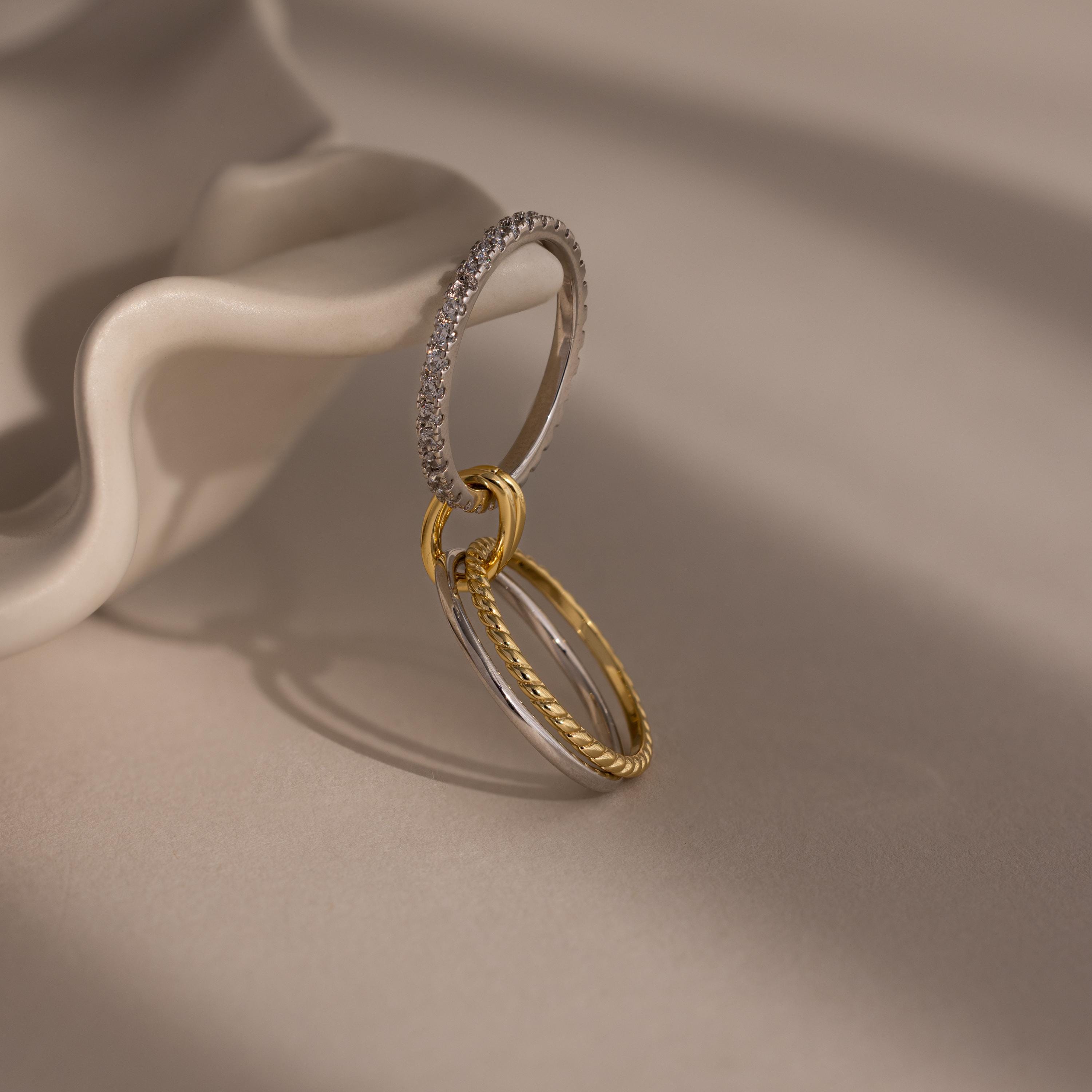 Three elegant rings from the Pave Mixed Metal Linked Ring Set, beautifully interlinked in gold and silver tones, rest on a smooth, wavy ceramic surface.