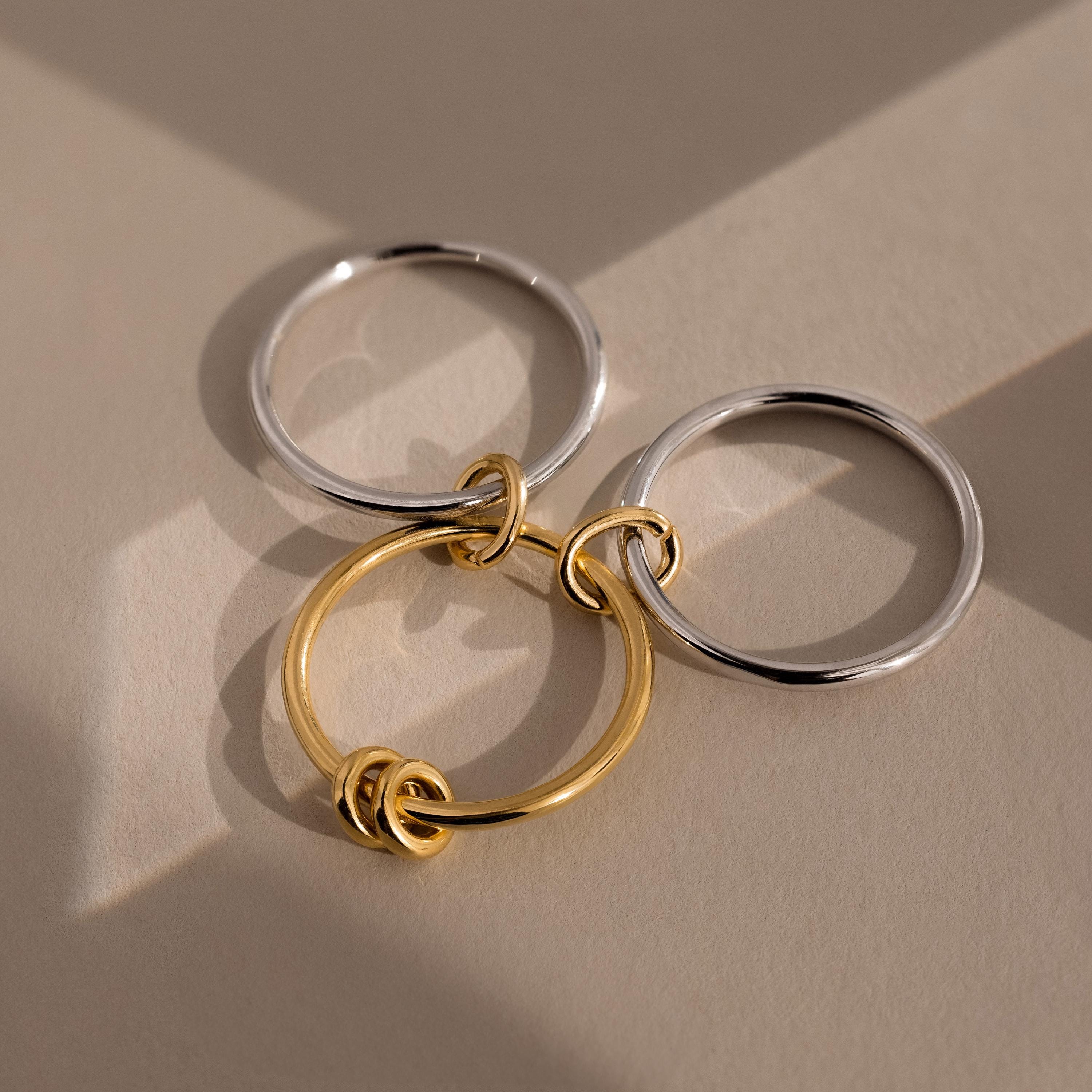 The Mixed Metal Linked Ring Set features three interlocking metal rings—two silver, one gold—that create a stylish accessory, casting soft shadows on a beige surface.
