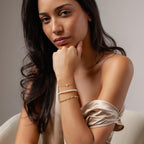 Woman in a satin off-shoulder top, wearing delicate jewelry including a gold chain bracelet and the Rounded Pearl Flower Bracelet, sits with her hand under her chin.