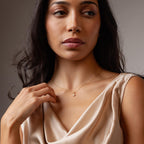 A woman in a cream dress gently touches the Rounded Pearl Flower Necklace in 18K Gold, gazing thoughtfully to the side.