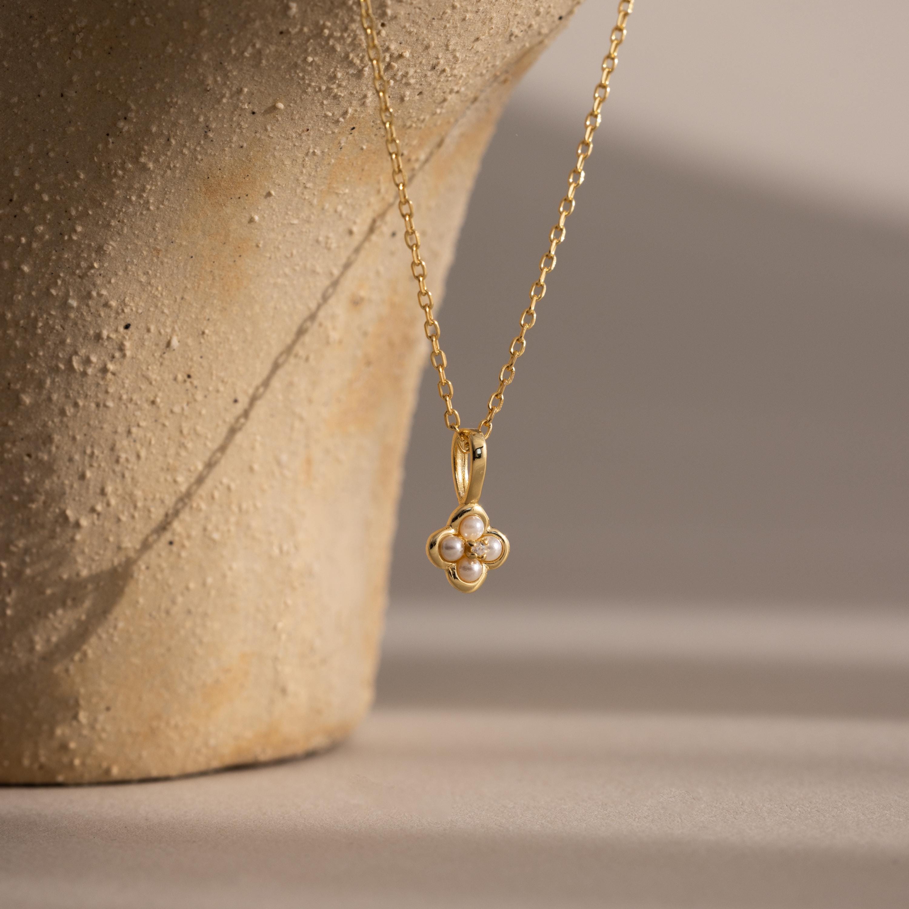 The Rounded Pearl Flower Necklace, featuring a gold chain and clover-shaped pendant with four pearls, is displayed elegantly in front of a textured vase, showcasing its refined floral charm.