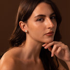 A woman with long brown hair and neutral makeup wears the Pearl Bezel Studs in 18K Gold, posing gracefully against a brown background—an elegant choice for everyday style.