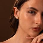 A woman with brown hair, wearing Pearl Bezel Studs, gazes at the camera with her hand near her face, highlighting the elegant classic jewelry.