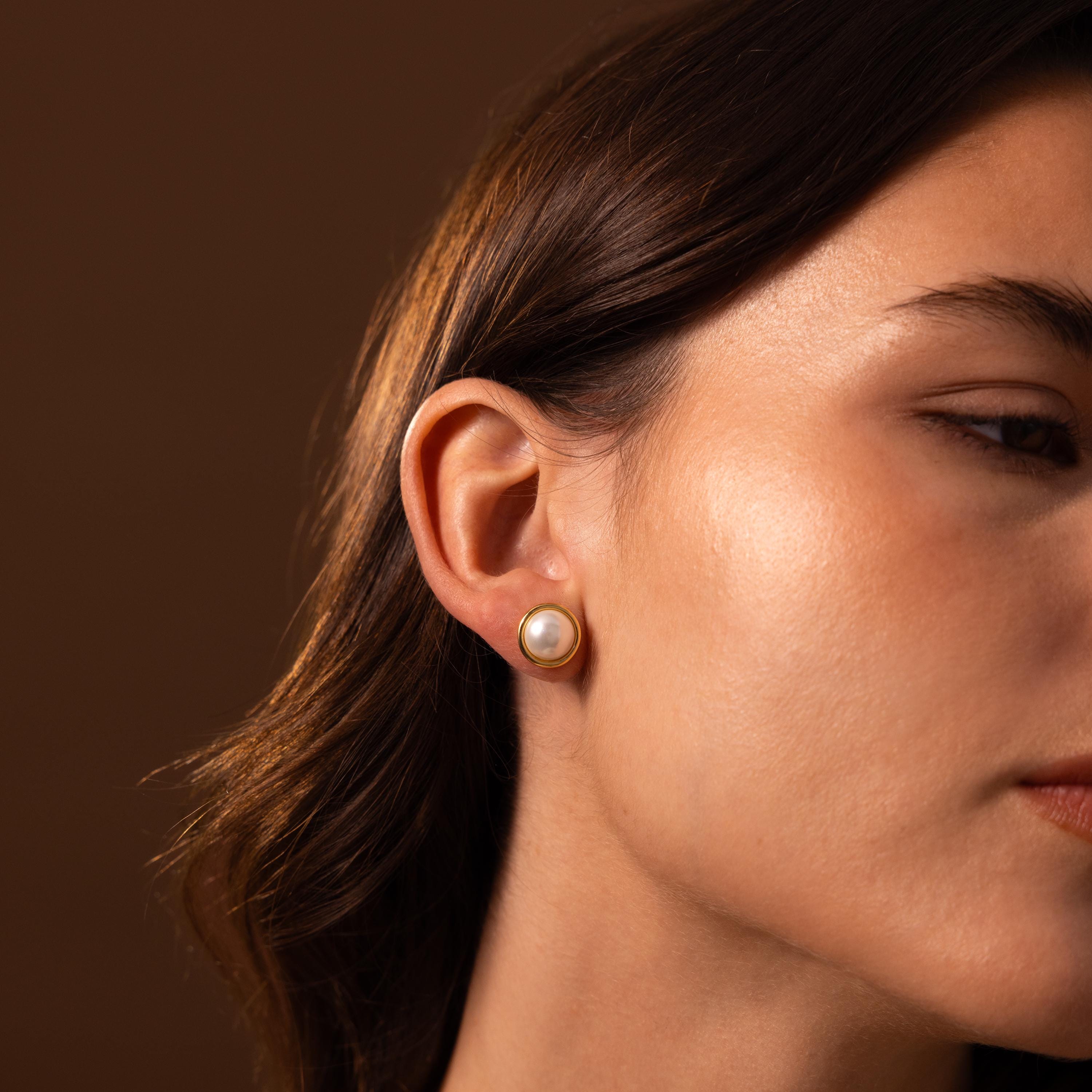 A close-up of a woman's ear adorned with Pearl Bezel Studs highlights the classic and timeless style of this elegant jewelry against a warm brown background.