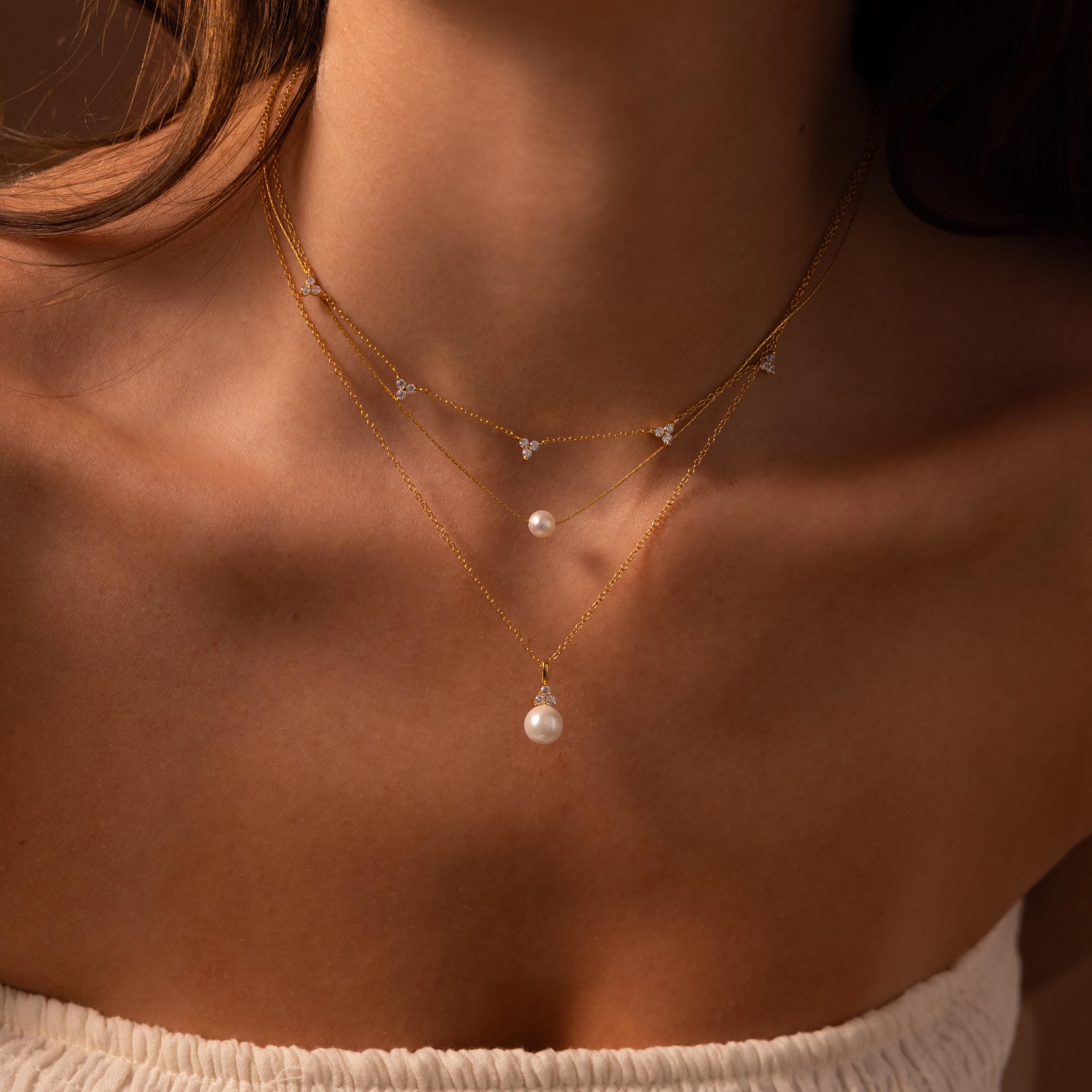 A woman showcases timeless jewelry: layered gold necklaces with pearls and crystals, featuring the captivating Diamond Halo Pearl Necklace, visible from shoulders to collarbone.