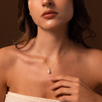 A woman touches her collarbone while wearing the Diamond Halo Pearl Necklace, a classic jewelry piece, against a brown background.