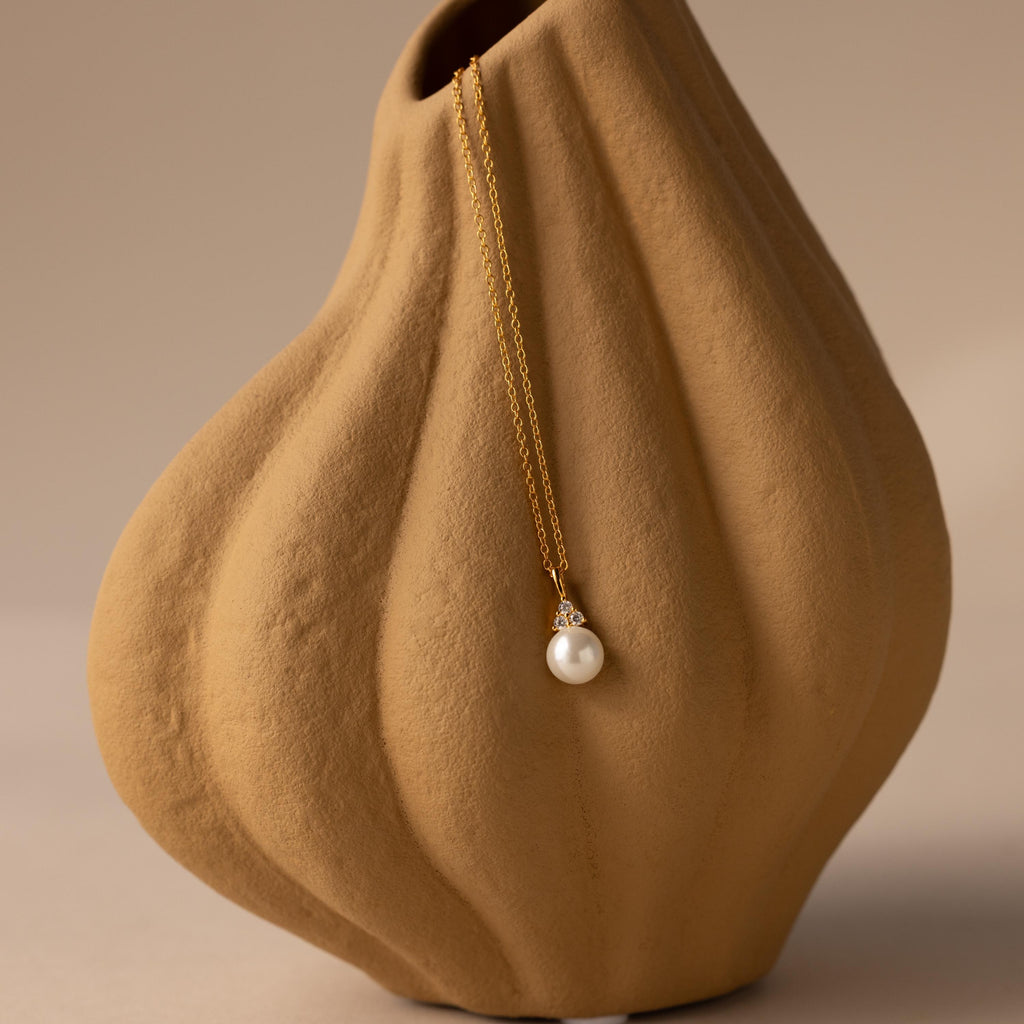 The Diamond Halo Pearl Necklace in 18K Gold, featuring a luminous pearl pendant, is elegantly displayed on a textured beige ceramic vase.