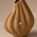 The Diamond Halo Pearl Necklace in 18K Gold, featuring a luminous pearl pendant, is elegantly displayed on a textured beige ceramic vase.