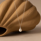 The Diamond Halo Pearl Necklace is elegantly showcased in front of a brown seashell, emphasizing the timeless beauty and sophistication of this stunning jewelry piece.