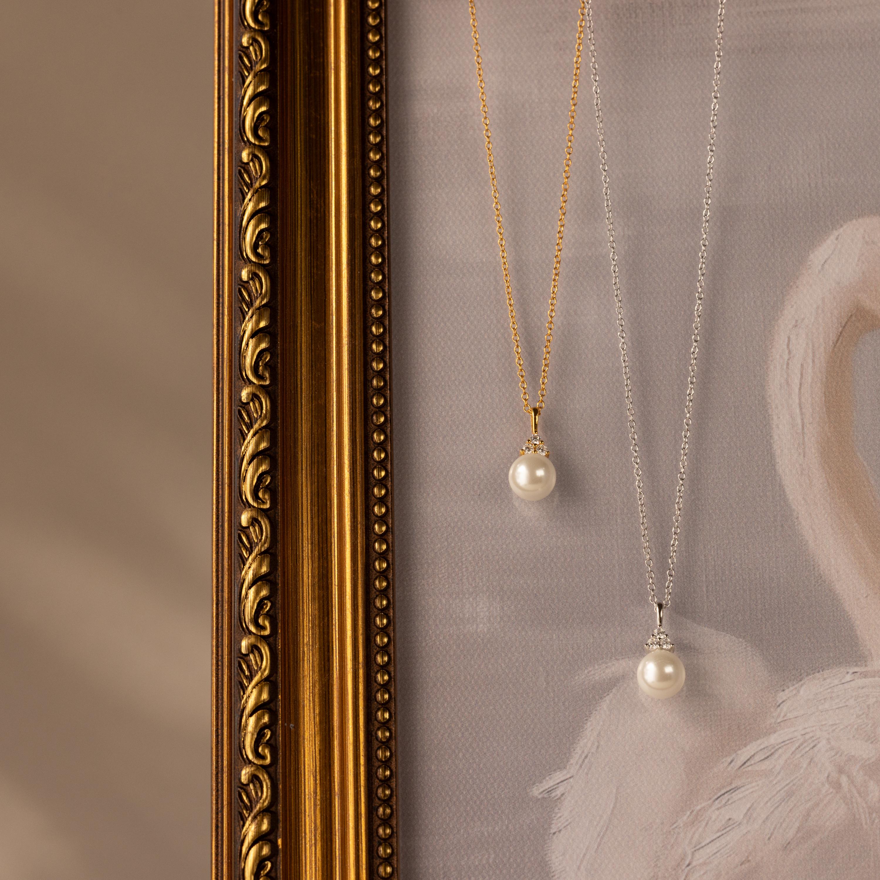 The Diamond Halo Pearl Necklace hangs gracefully over a framed artwork featuring a golden ornate edge and a white swan illustration.