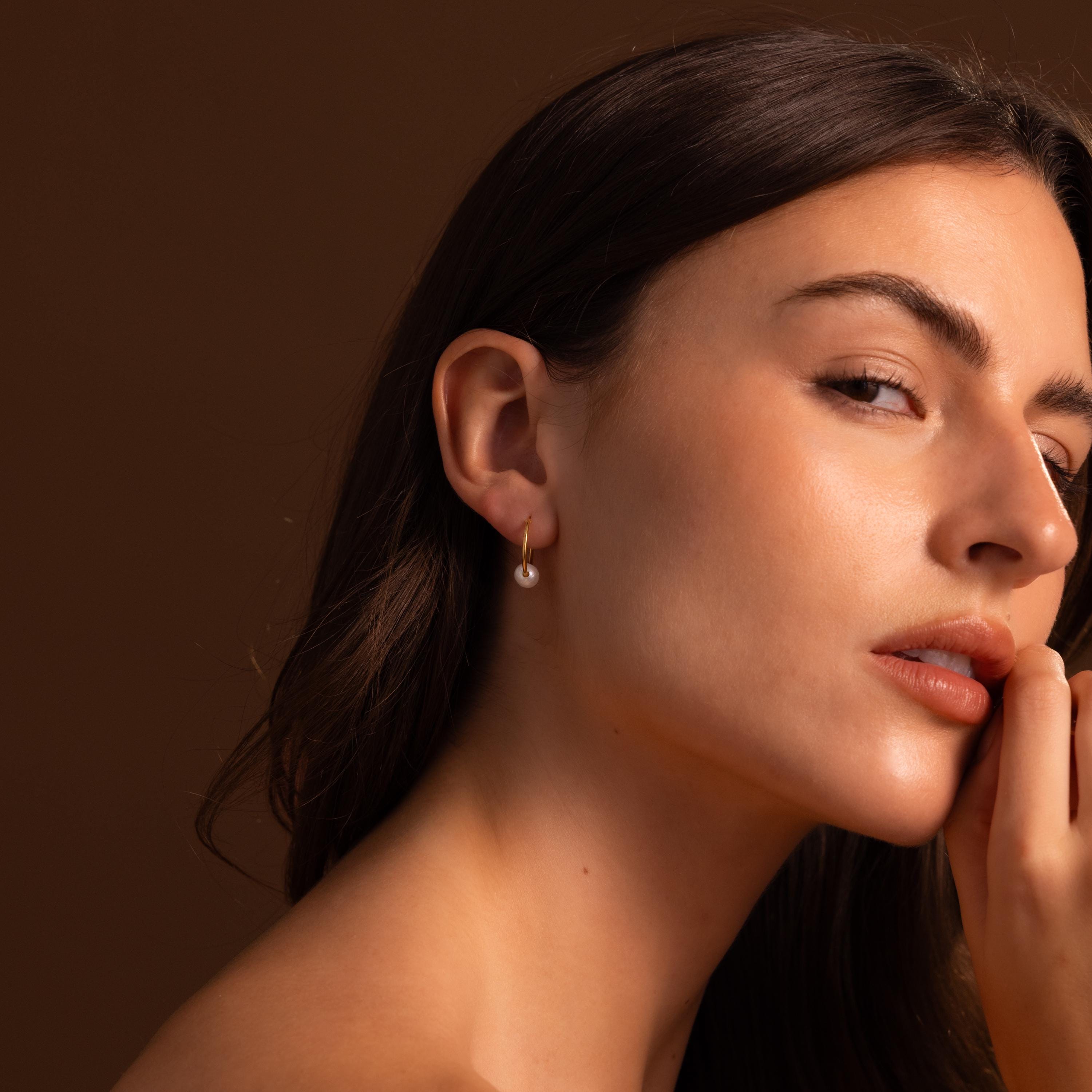 A woman with long brown hair wears Round Pearl Hoops, posing against a brown background.