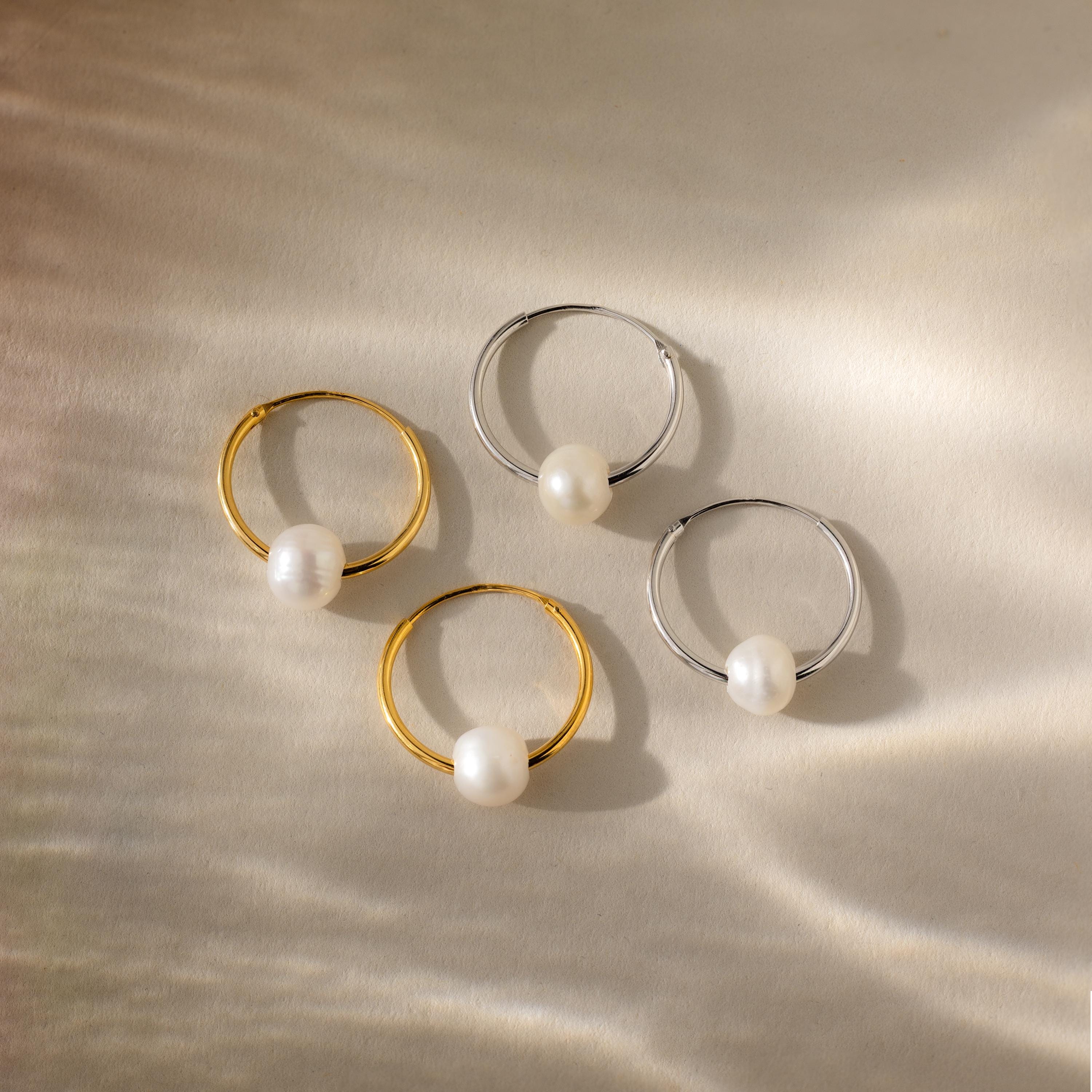 Two gold and two silver Round Pearl Hoops, each adorned with a single pearl, arranged on a softly lit textured surface—an elegant display of timeless jewelry.