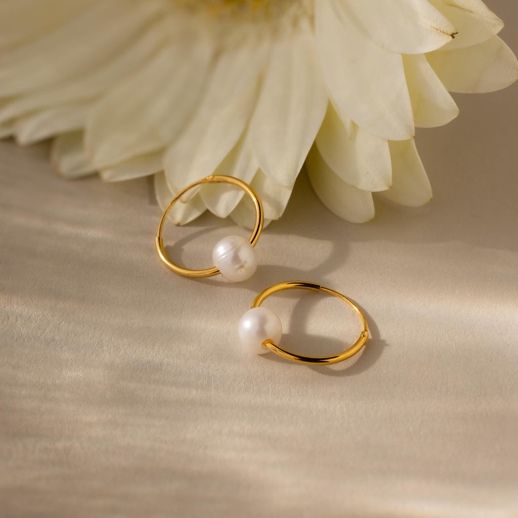Round Pearl Hoops with delicate accents rest on a light surface near a white flower, embodying timeless jewelry elegance.
