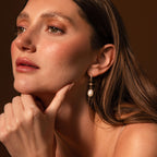 Woman with long brown hair wears North Star Pearl Hoops in 18K Gold, gazing thoughtfully to the side with her hand on her chin.