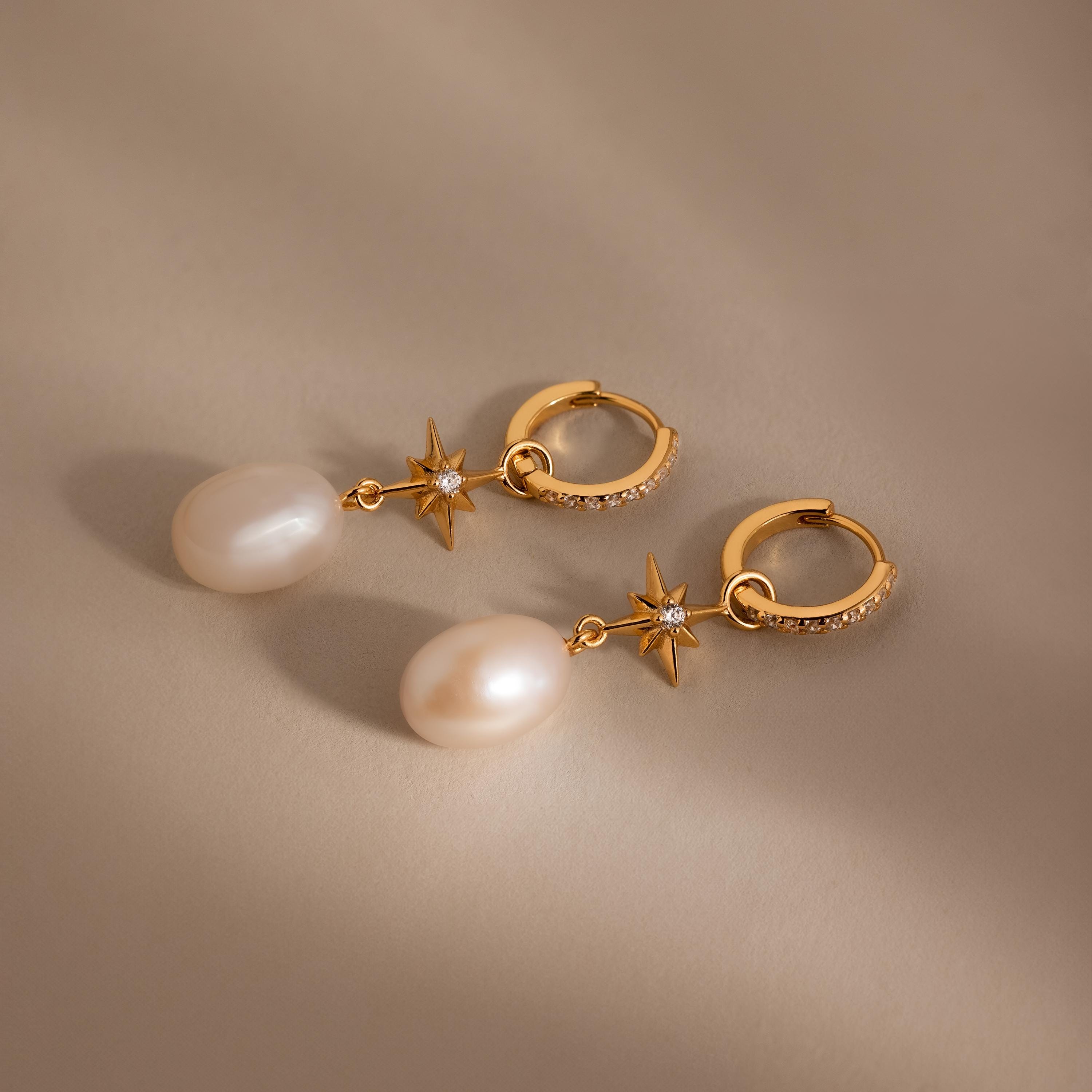 North Star Pearl Hoops in 18K Gold—gold hoop earrings with pearl drops and star-shaped accents inspired by celestial jewelry—shown on a neutral beige background.