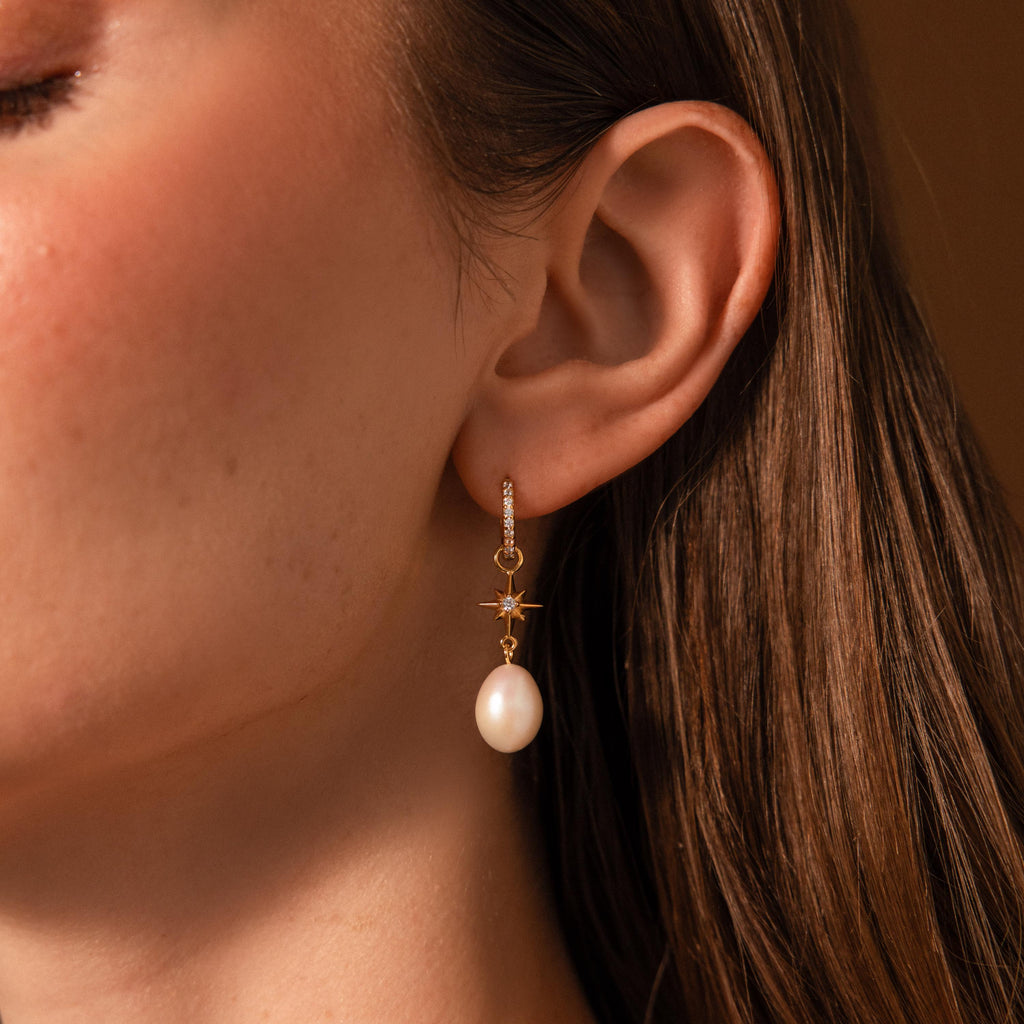 Close-up of a woman's ear wearing the North Star Pearl Hoops, gold earrings featuring a star charm and dangling white pearl.