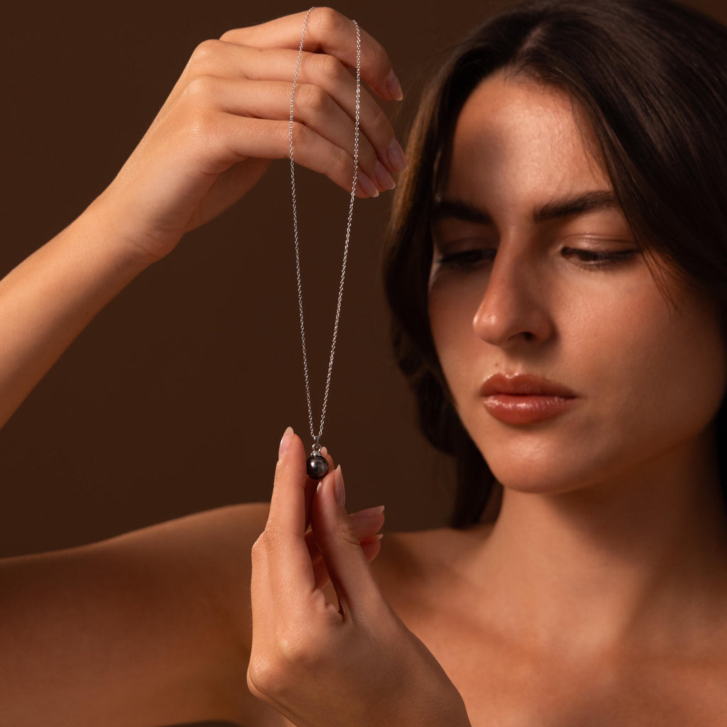 A woman holds up and admires the elegant Midnight Pearl Necklace in Sterling Silver, featuring a dark pendant, set against a brown background.