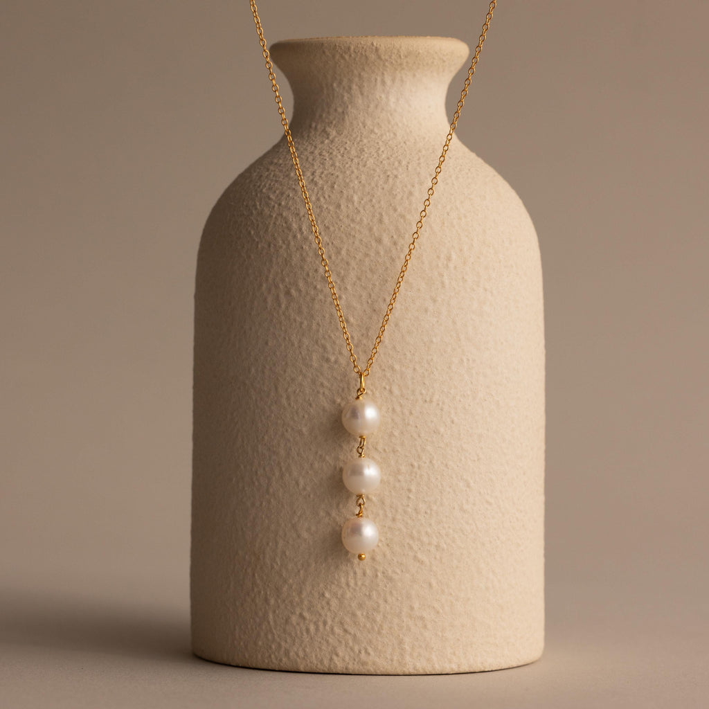 The Pearl Cascade Necklace in 18K Gold features three stacked pearls, elegantly draped over a textured beige vase—an ideal piece for everyday wear against a neutral backdrop.