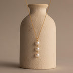 The Pearl Cascade Necklace in 18K Gold features three stacked pearls, elegantly draped over a textured beige vase—an ideal piece for everyday wear against a neutral backdrop.