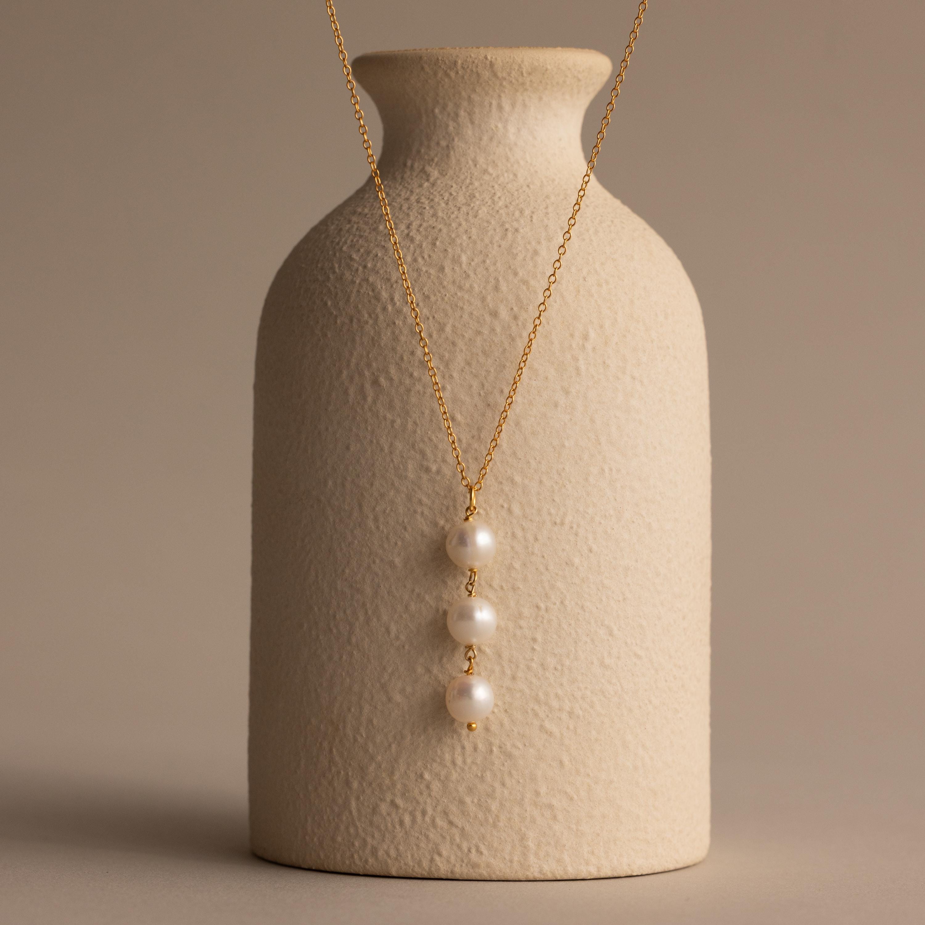The Pearl Cascade Necklace in 18K Gold features three stacked pearls, elegantly draped over a textured beige vase—an ideal piece for everyday wear against a neutral backdrop.
