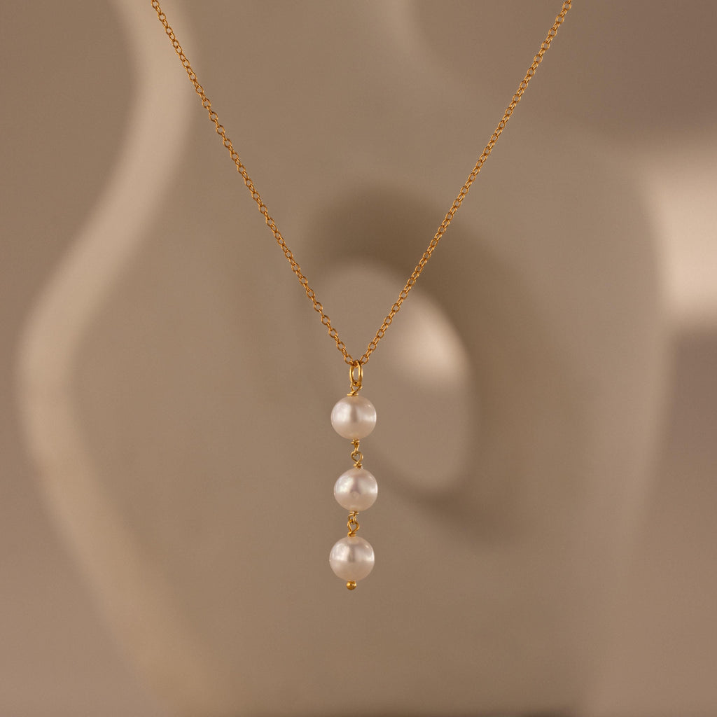 The Pearl Cascade Necklace features three luminous pearls in a vertical drop on a gold chain, set against a soft beige abstract background.