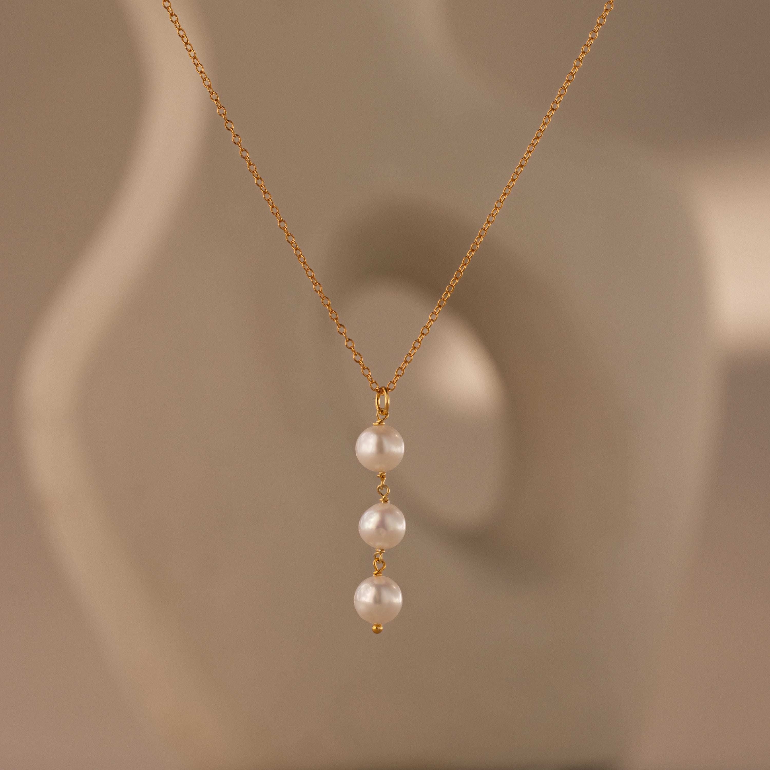 The Pearl Cascade Necklace features three luminous pearls in a vertical drop on a gold chain, set against a soft beige abstract background.